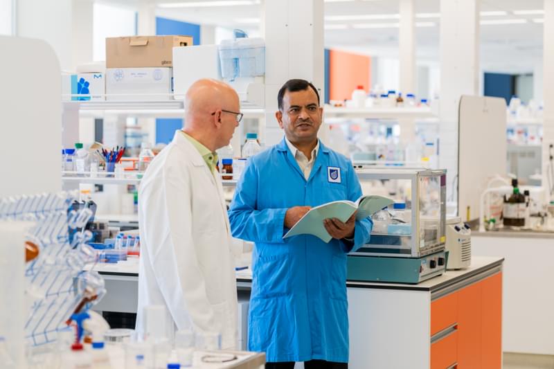 UniSA Centre for Pharmaceutical Innovation Directors stand with lab coast, in a lab talking to each other. One holds a book. 