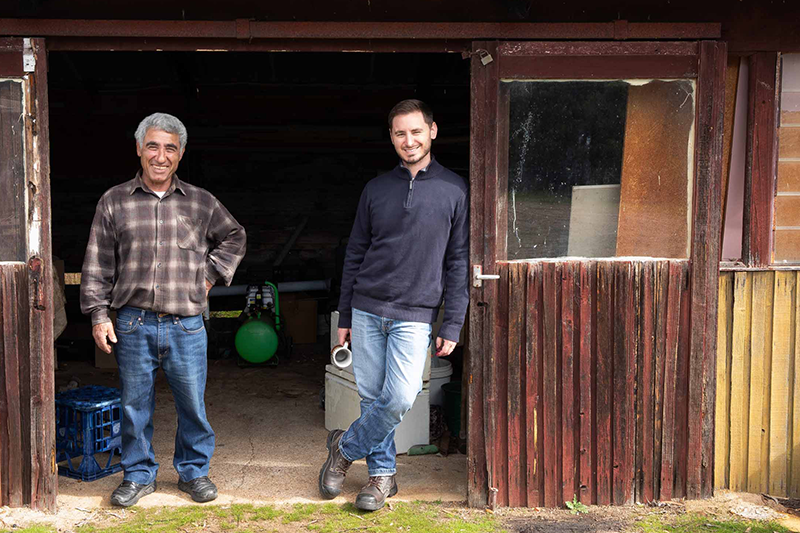 Two olive farmers smile, facing the camera. They are framed in the wide doorway of a rundown looking iron and wood shed. One man holds a china mug.png