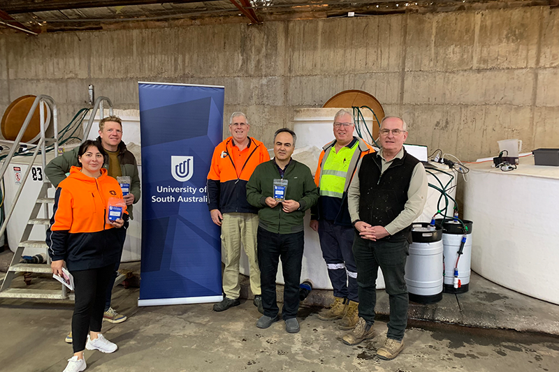 UniSA researchers and collaborators stand in an industrial warehouse in front of a blue UniSA pull-up banner. Some hold little plastic sachets of olives. .png