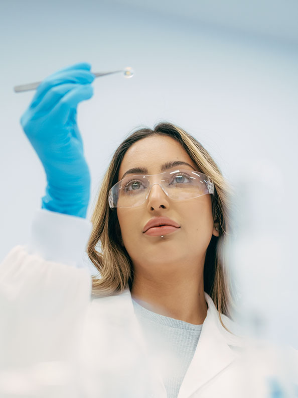 A researchers in white lab coat examines a small specimen, perhaps a mineral, between some silver tweezers. 