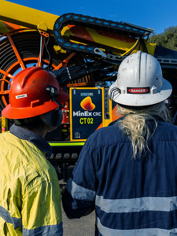 Two people wearing hi-vis uniforms and hard hats face a large yellow drilling machine. Their backs are to the camera