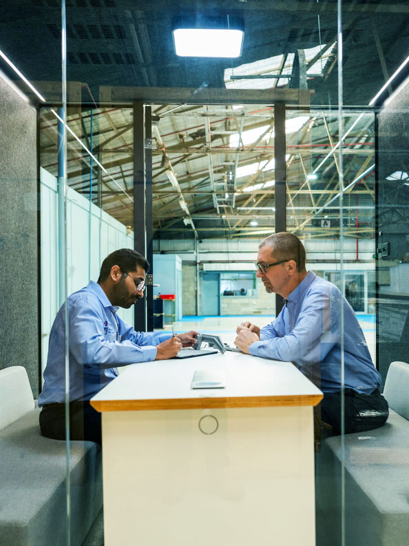 Two men dressed in light blue shirts sit inside a glass room, facing each other. There is a wooden table between them. The glass room is situated in a larger facility, which appears to be industrial in nature.